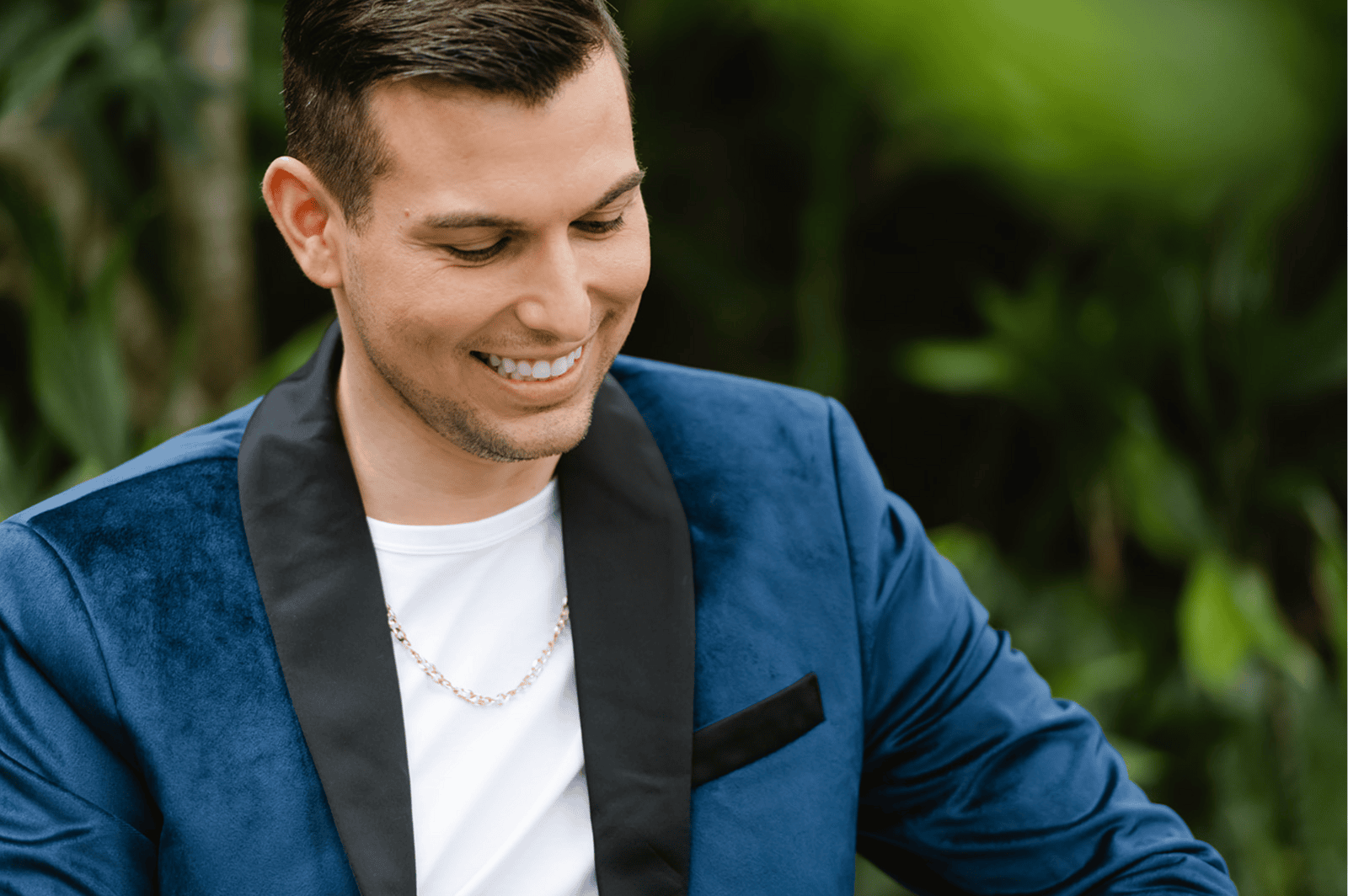 Matt Fraser is smiling, wearing a blue velvet suit jacket, white shirt, and gold chain, standing outdoors with greenery in the background.